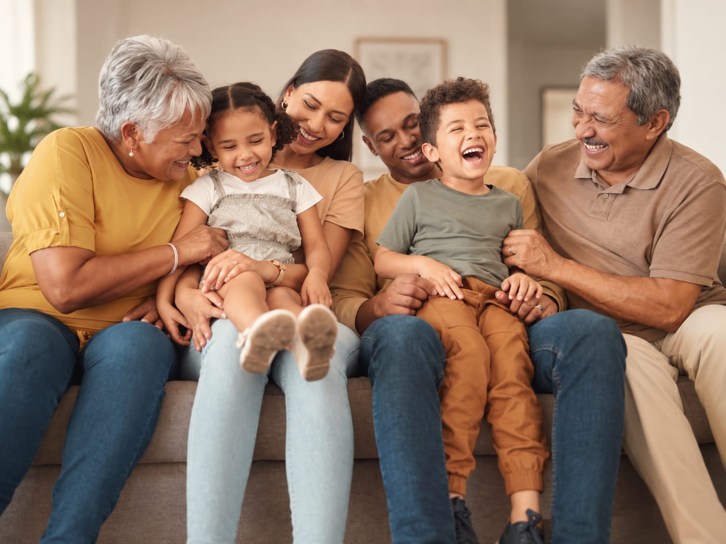 Three generations laughing together on a sofa: grandparents, parents, and two young children on their laps.