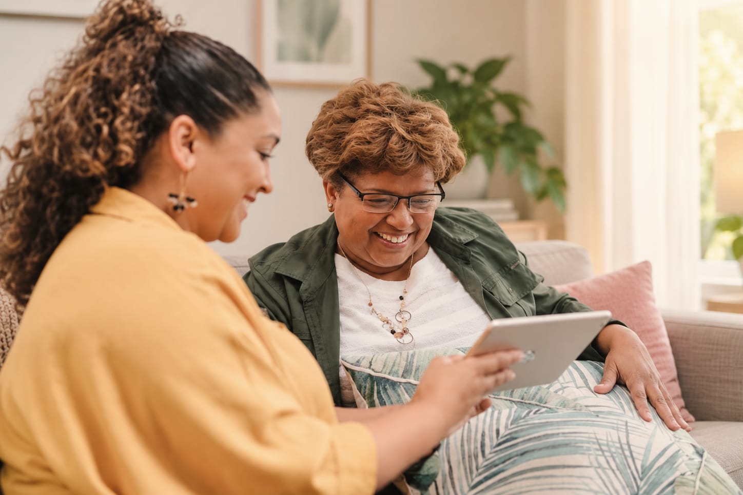 A woman with short curly hair and glasses laughs as her adult daughter shows her something on a tablet on the sofa.