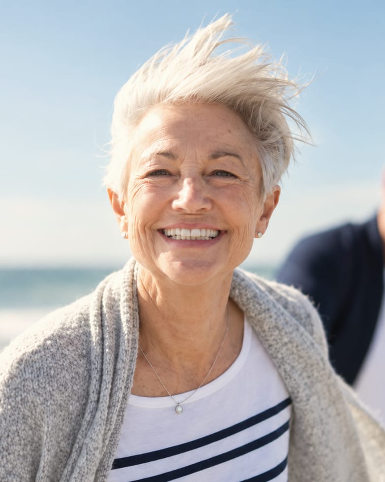 A woman with short white hair laughs on a windy beach in a striped top and a grey cardigan, a man walking behind her.