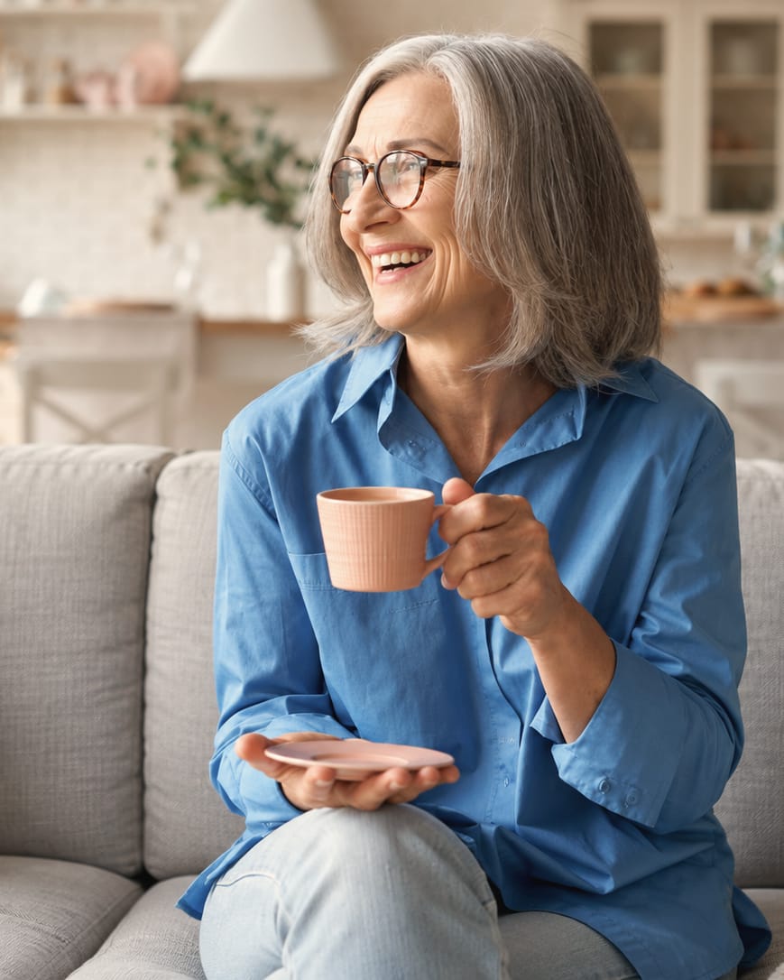 A woman in a blue shirt laughs on a sofa, holding a pink cup and saucer.