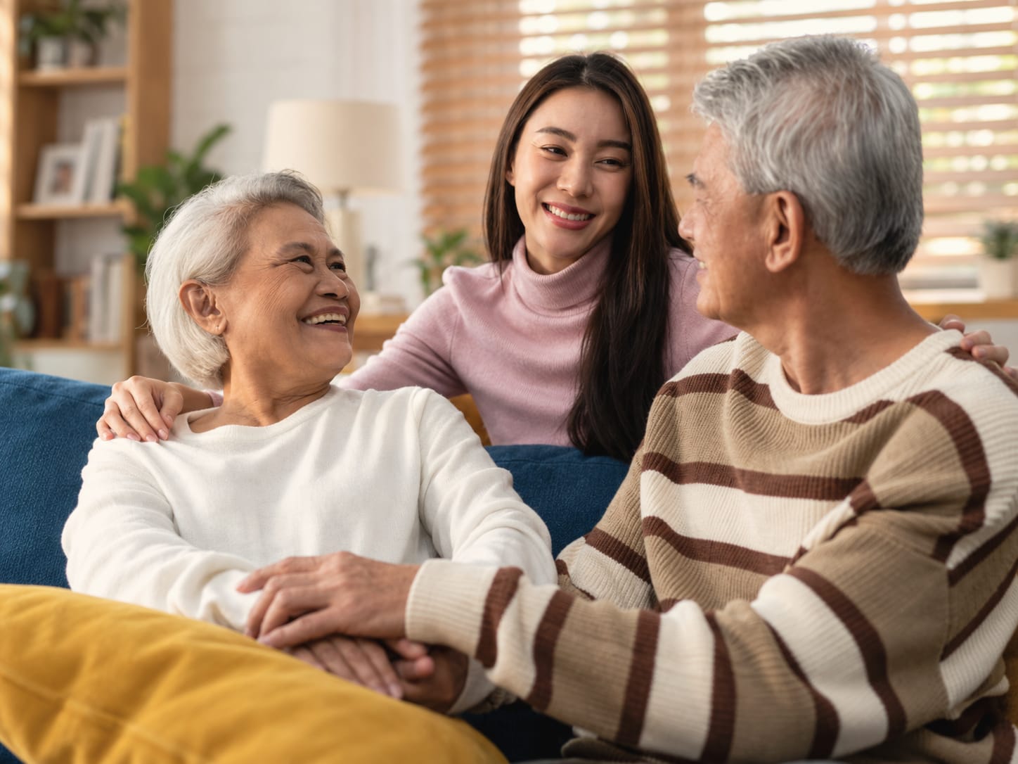 A younger woman leans between an older woman and an older man on a sofa, all three laughing, their hands together.