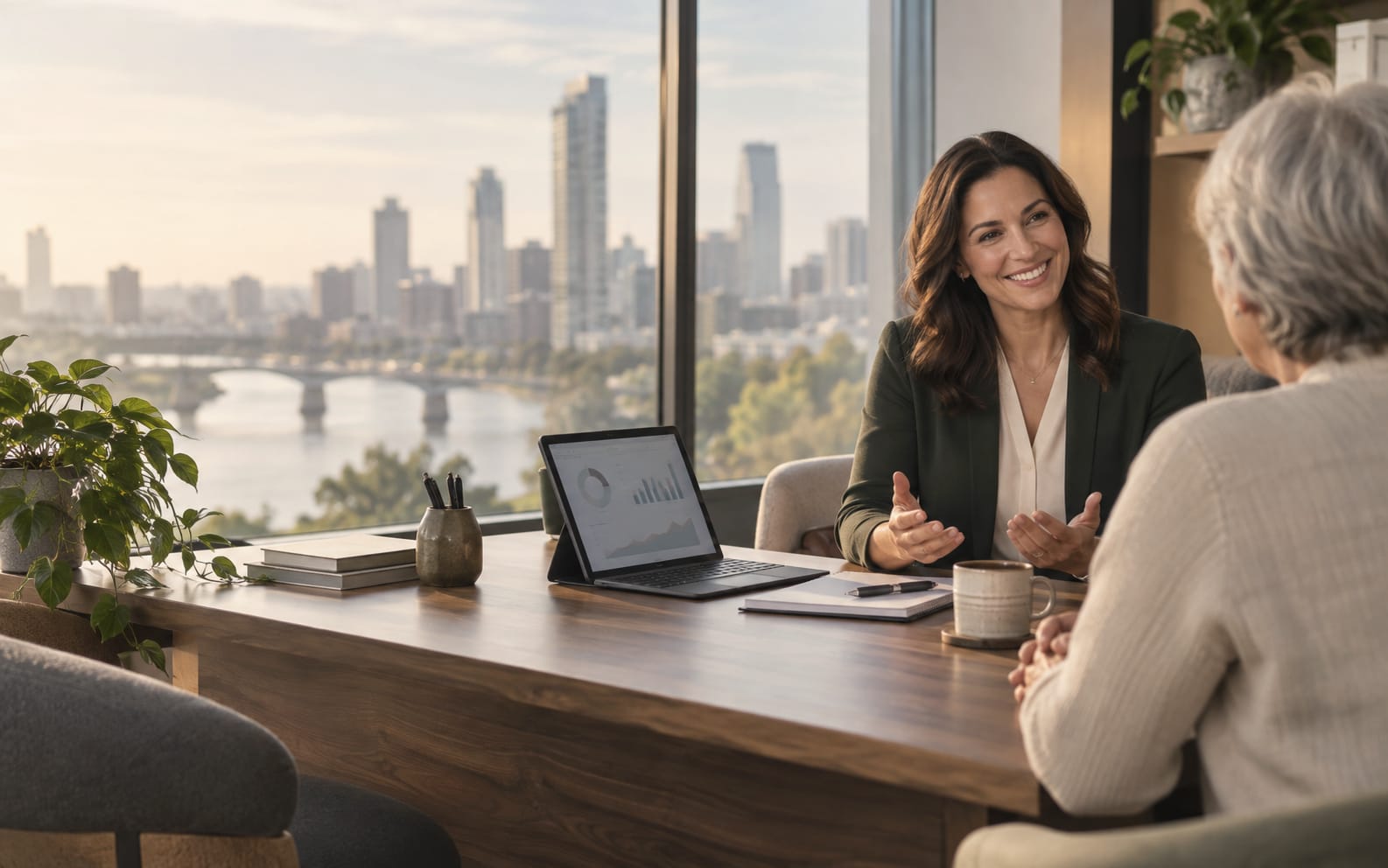 A woman in a dark green blazer talks across a desk with an older woman, a tablet and notebook between them and a city skyline and river through the window.