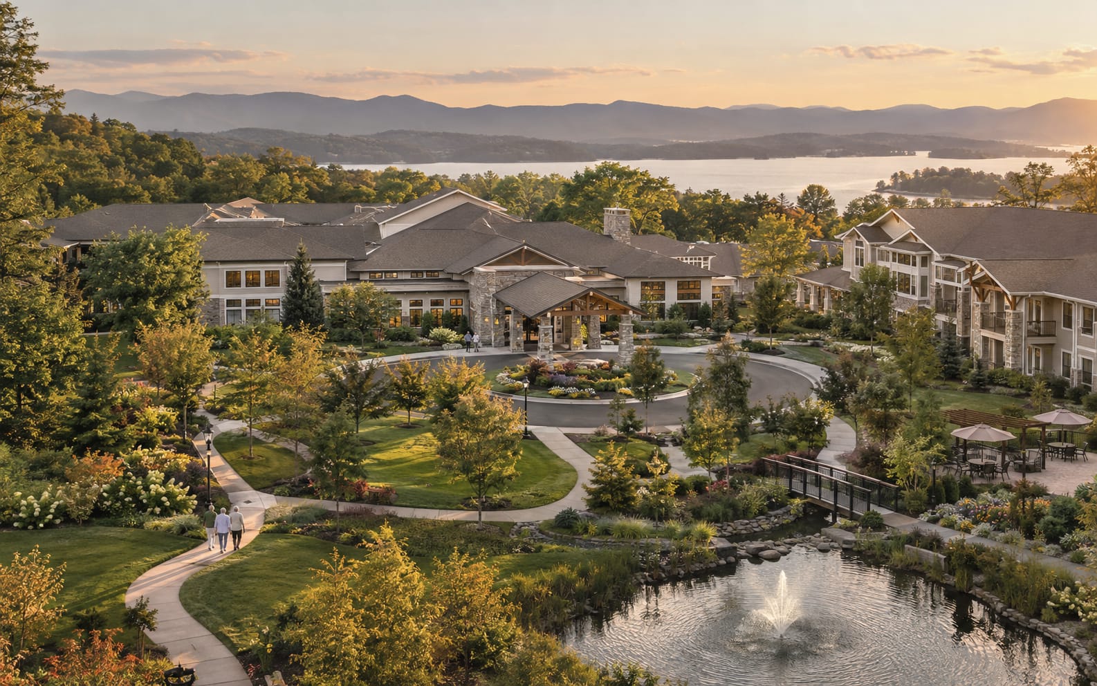 A senior living campus seen from above at sunset: a circular drive, a pond with a fountain, and a lake and mountains behind.