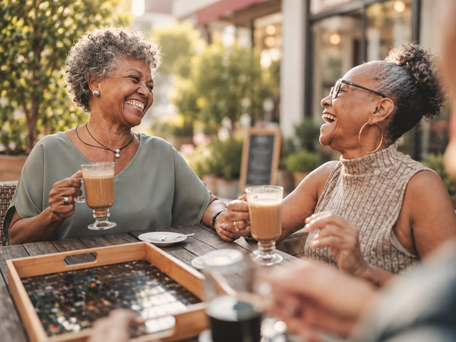 Two women laughing together over glass mugs of coffee at an outdoor cafe table.